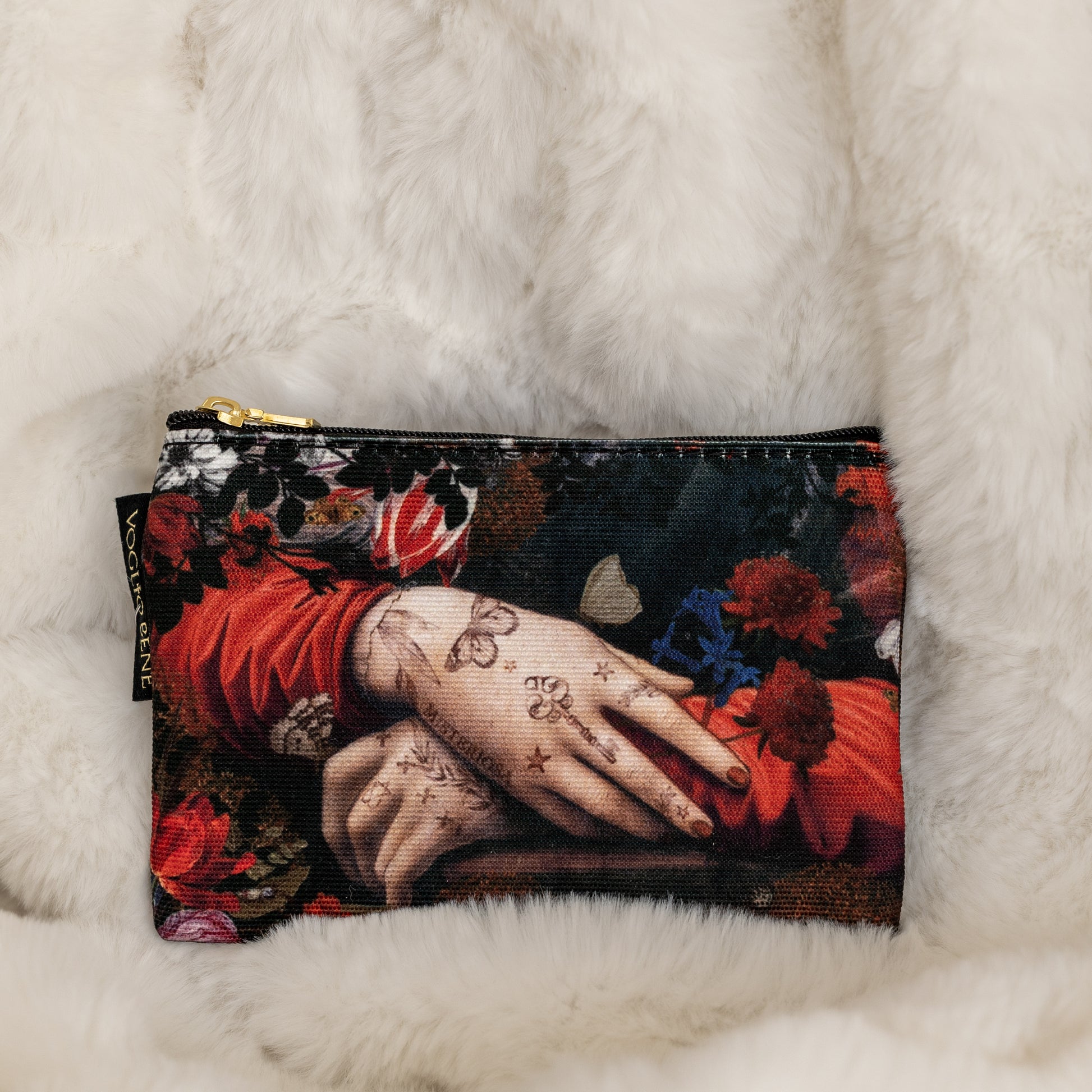 Pieni kukkakuvioinen kangaslaukku tai lompakko, jonka kuvituksessa tatuoitu käsi lepää punaisten ja sinisten kukkien keskellä. - Small floral-patterned fabric pouch featuring a tattooed hand resting among red and blue flowers.