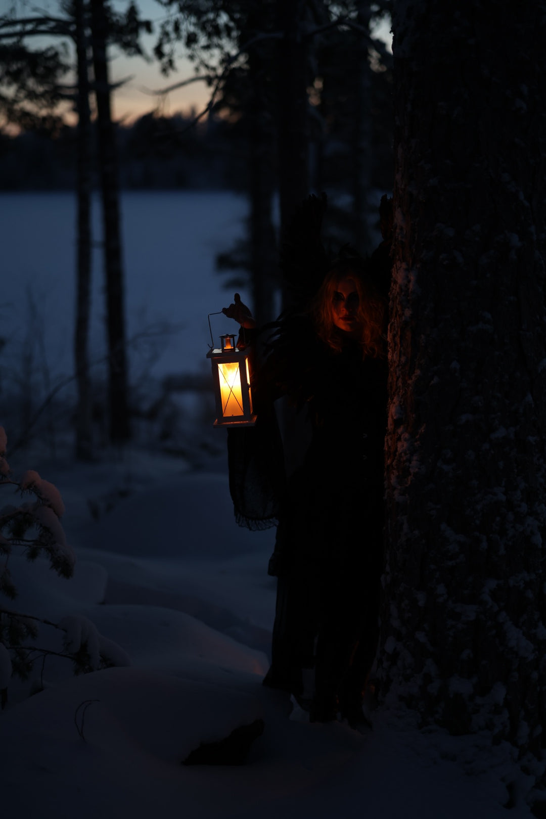 Tummaan asuun pukeutunut hahmo pitää palavaa lyhtyä puun vieressä lumisessa metsässä.
