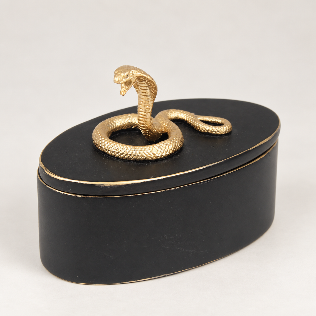 Musta ovaali rasia, jonka irrotettavan kannen päällä on kullanväriseksi maalattu kobra ja reunoissa kullanväriset korostukset. / Black oval trinket box with a gold-painted cobra on the removable lid and gold-coloured edging.