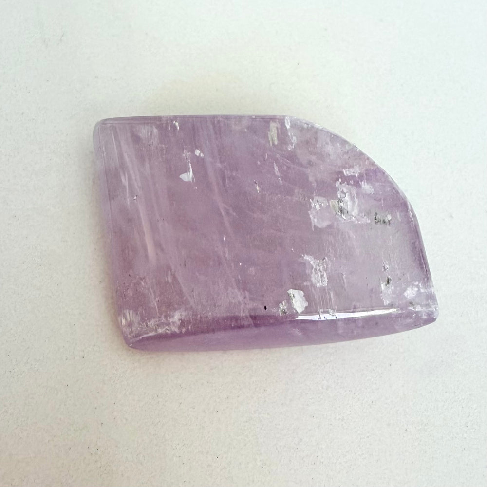 Suoraan päältä otettu kuva vaaleanvioletista, sileäksi hiotusta kunziittikivestä. Kivi on laakea, epäsäännöllisen neliskulmainen ja sen pinta on osittain matta, osittain kiiltävä. Kiven sisällä näkyy lukuisia luonnollisia, valoa heijastavia säröjä ja sulkeumia, jotka luovat sille ominaisen ulkonäön.