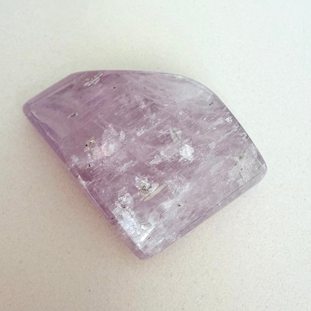 Yläviistosta otettu kuva vaaleanvioletista, puoliläpinäkyvästä ja kiillotetusta kunziittikivestä valkoisella taustalla. Kivi on muodoltaan epäsäännöllisen neliskulmainen ja sen pinta on täynnä luonnollisia, hohtavia halkeamia. Valo heijastuu kiven pinnasta tuoden esiin sen sisäisen rakenteen.