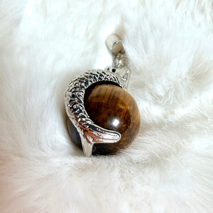 FI: Hopeanvärinen merenneito-riipus, joka pitää kullanruskeaa tiikerinsilmä-kivipalloa.
EN: Silver-coloured mermaid pendant holding a golden-brown tiger’s eye stone sphere.