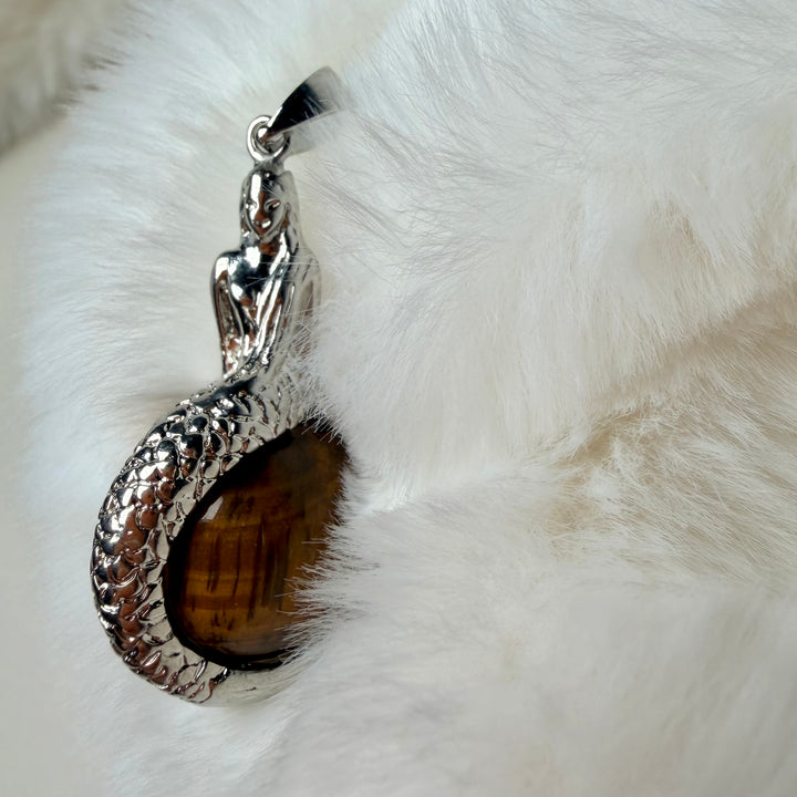 FI: Hopeanvärinen merenneito-riipus, joka pitää kullanruskeaa tiikerinsilmä-kivipalloa.
EN: Silver-coloured mermaid pendant holding a golden-brown tiger’s eye stone sphere.