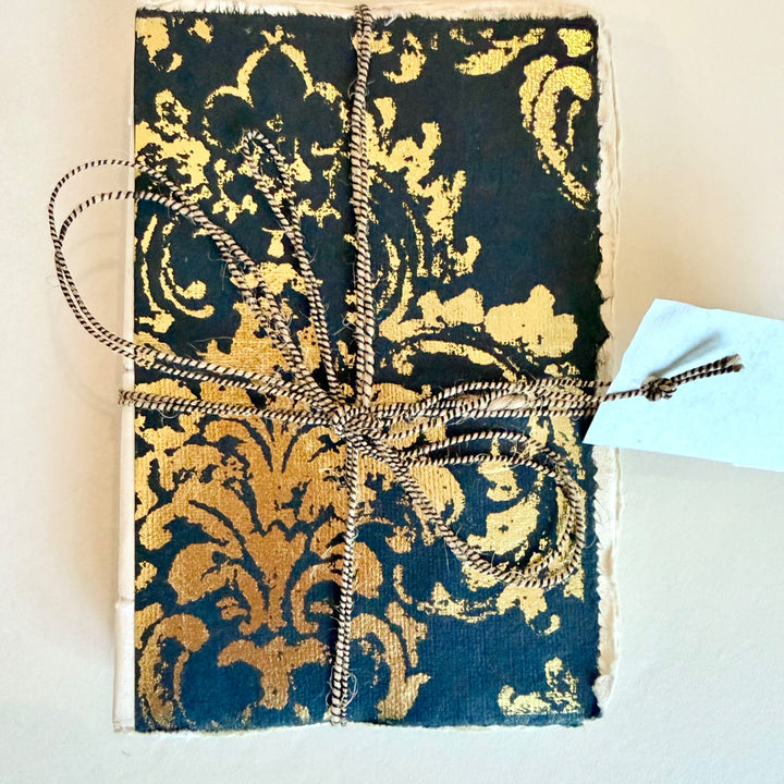 Grand Siècle -muistikirja, musta kansi kullanvärisellä koristekuviolla ja nyöri. / Grand Siècle notebook with a black cover, gold-coloured ornament and cord.