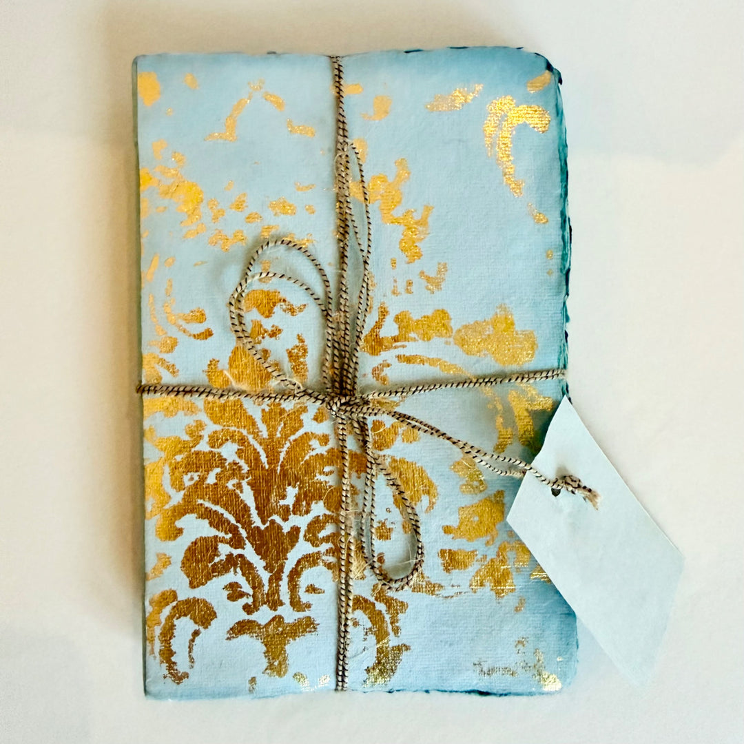 Grand Siècle -muistikirja, vaaleansininen kansi kullanvärisellä koristekuviolla ja nyöri. / Grand Siècle notebook with a light blue cover, gold-coloured ornament and cord.