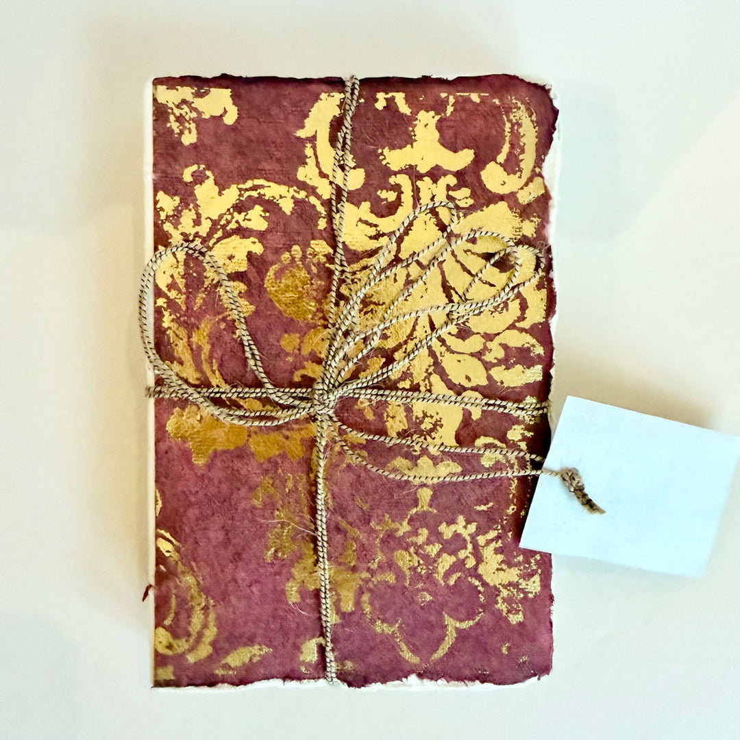 Grand Siècle -muistikirja, viininpunainen kansi kullanvärisellä koristekuviolla ja nyöri. / Grand Siècle notebook with a burgundy cover, gold-coloured ornament and cord.