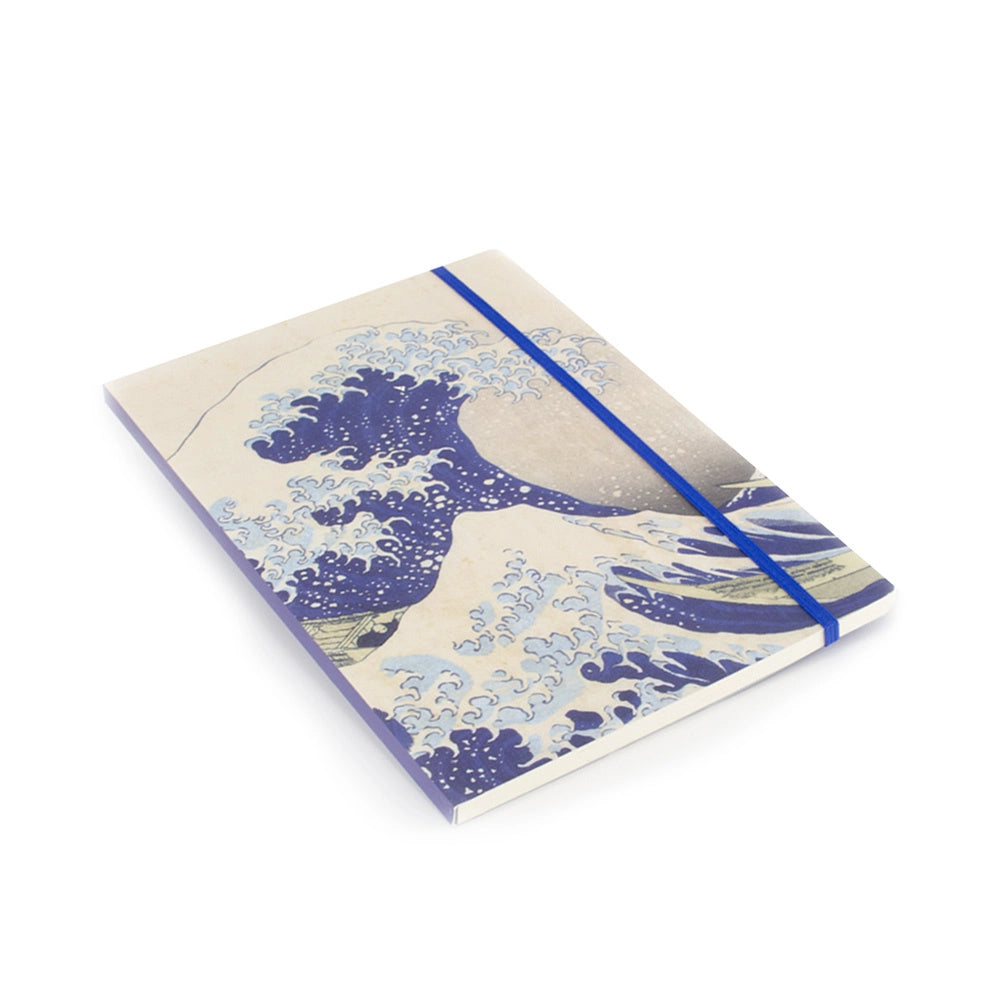 Hokusain Suuri aalto -muistikirja viistosta kuvattuna, näkyvissä kansikuva, sininen kuminauha ja sivujen paksuus. – Angled view of the Hokusai Great Wave notebook showing the cover illustration, blue elastic closure and page edges.