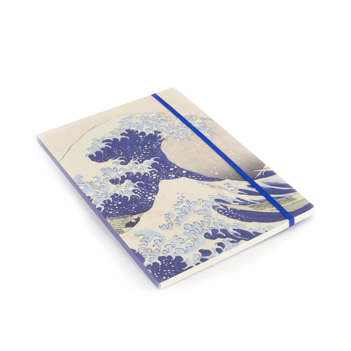 Hokusain Suuri aalto -muistikirja viistosta kuvattuna, näkyvissä kansikuva, sininen kuminauha ja sivujen paksuus. – Angled view of the Hokusai Great Wave notebook showing the cover illustration, blue elastic closure and page edges.
