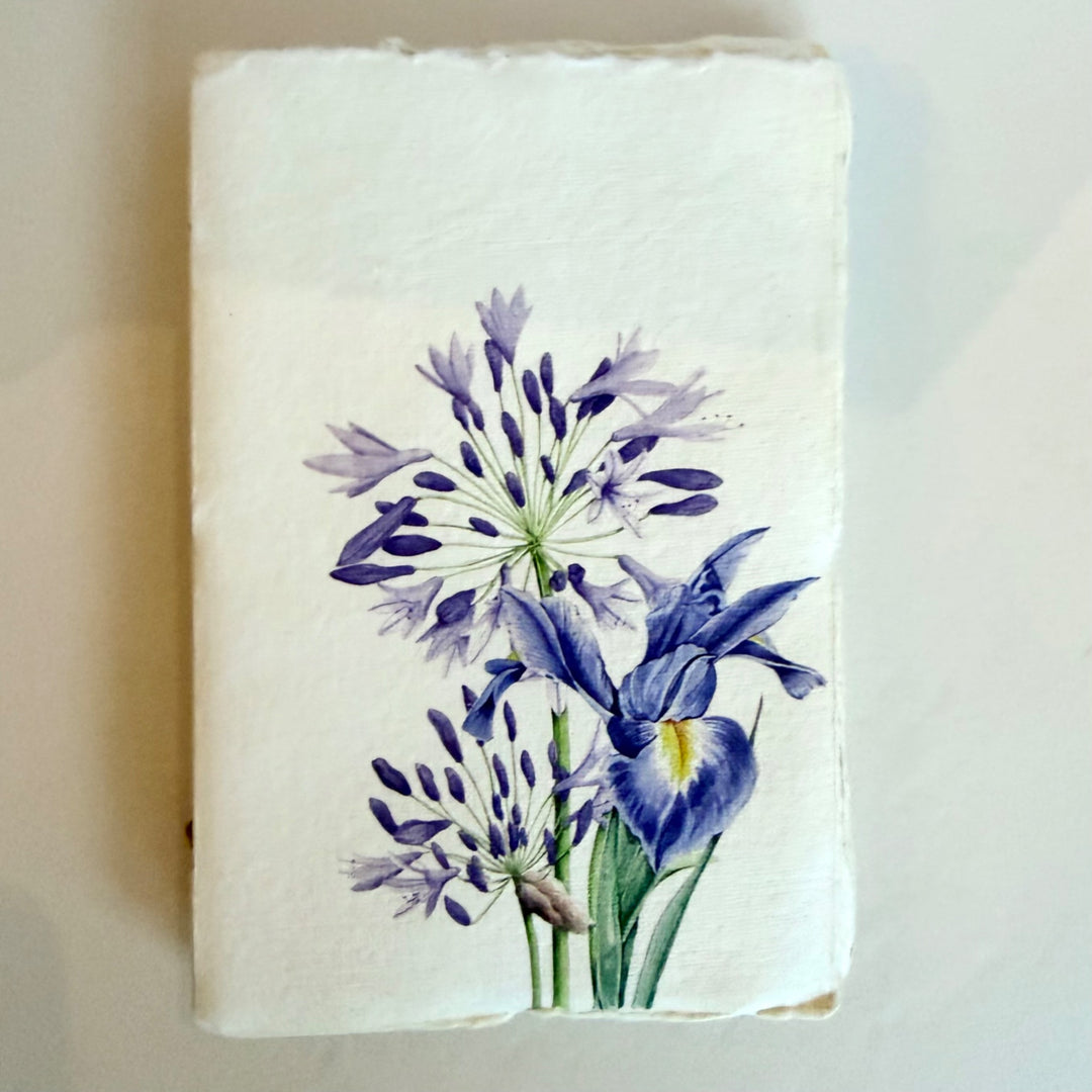 Vaalea käsintehty muistikirja, jonka kannessa on sininen iiris eli kurjenmiekka. / Cream handmade notebook with a blue iris on the cover.