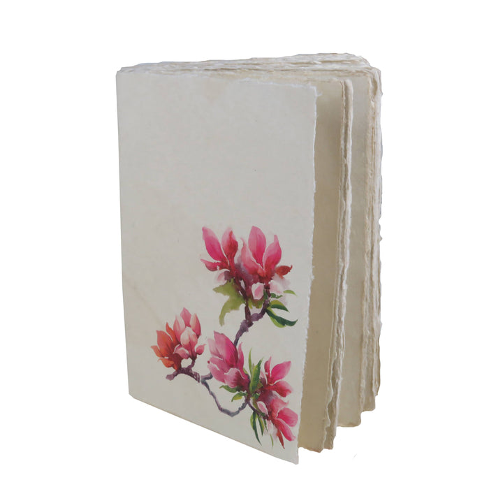Vaalea muistikirja, jonka kannessa on vaaleanpunaisia magnoliankukkia. / Cream notebook with pink magnolia flowers on the cover.