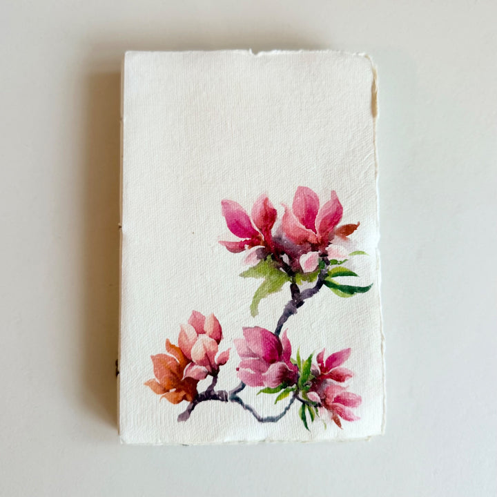 Vaalea käsintehty muistikirja, kuva-aiheena magnolia. / Cream handmade notebook with pink magnolia flowers on the cover.