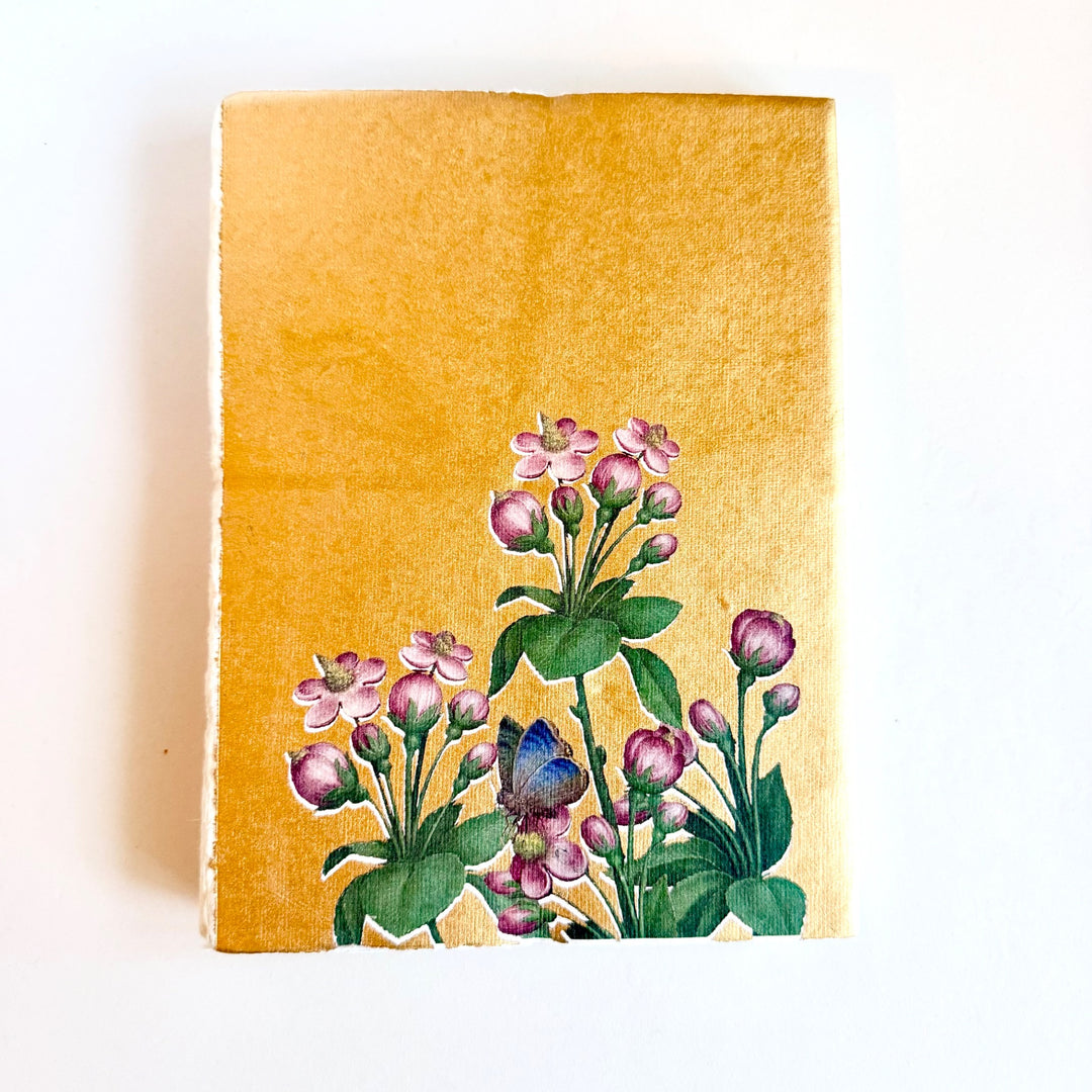Kultapohjainen muistikirja, jonka kannessa on vaaleanpunaisia kukkia ja sininen perhonen. / Gold-background notebook with pink flowers and a blue butterfly on the cover.