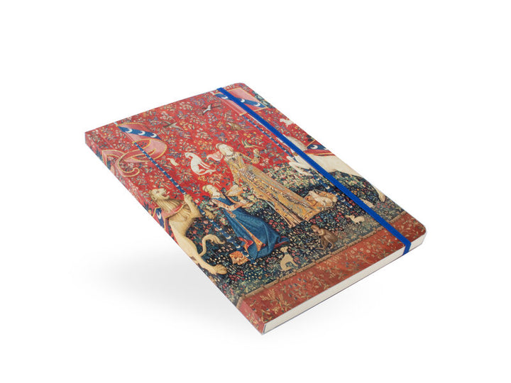 Nainen ja yksisarvinen -muistikirja viistosta kuvattuna, näkyvissä kuvakudosaiheinen kansi, sininen kuminauha ja sivujen paksuus. – Angled view of The Lady and the Unicorn notebook showing the tapestry cover, blue elastic closure and page edges.