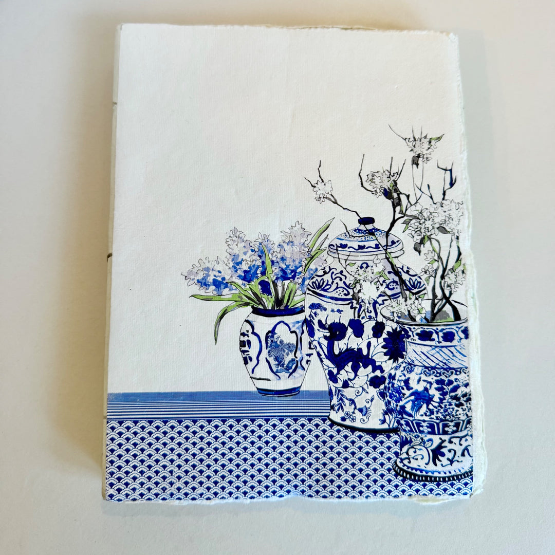 Sinivalkoinen muistikirja, jonka kannessa on koristeellisia maljakoita ja kukkia. / Blue and white notebook with decorative vases and flowers on the cover.