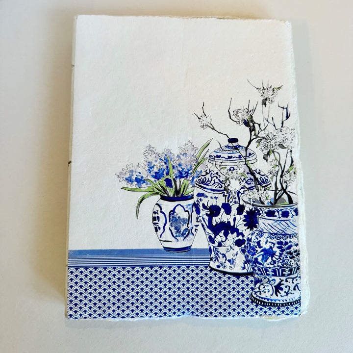 Sinivalkoinen muistikirja, jonka kannessa on koristeellisia maljakoita ja kukkia. / Blue and white notebook with decorative vases and flowers on the cover.
