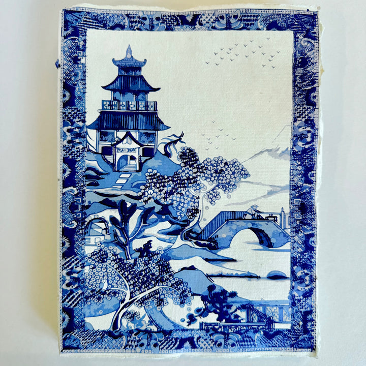 Sinivalkoinen muistikirja, jonka kannessa on puutarhamaisema, silta ja paviljonki. / Blue and white notebook with a garden landscape, bridge and pavilion.