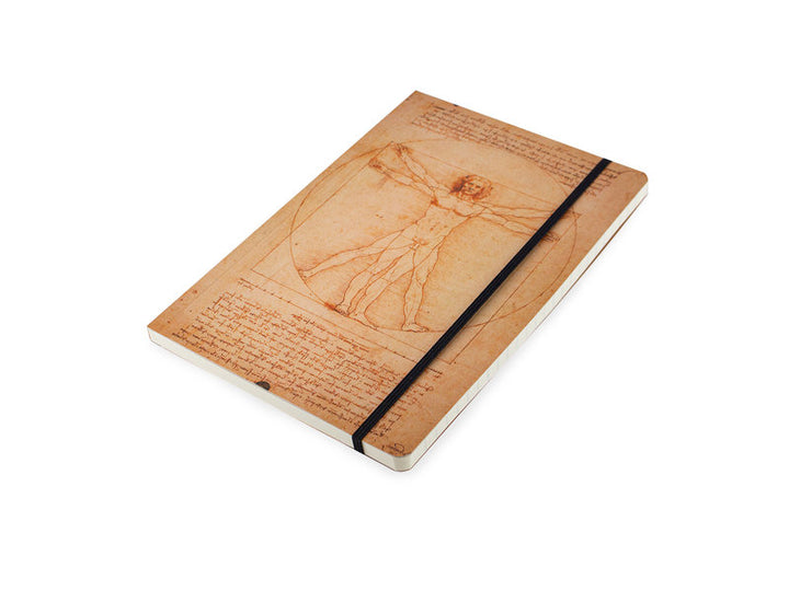 Pehmeäkantinen Vitruviuksen mies -muistikirja viistosta kuvattuna, näkyvissä kansikuva, musta kuminauha ja sivujen paksuus. – Angled view of the softcover Vitruvian Man notebook showing the cover illustration, black elastic closure and page edges.