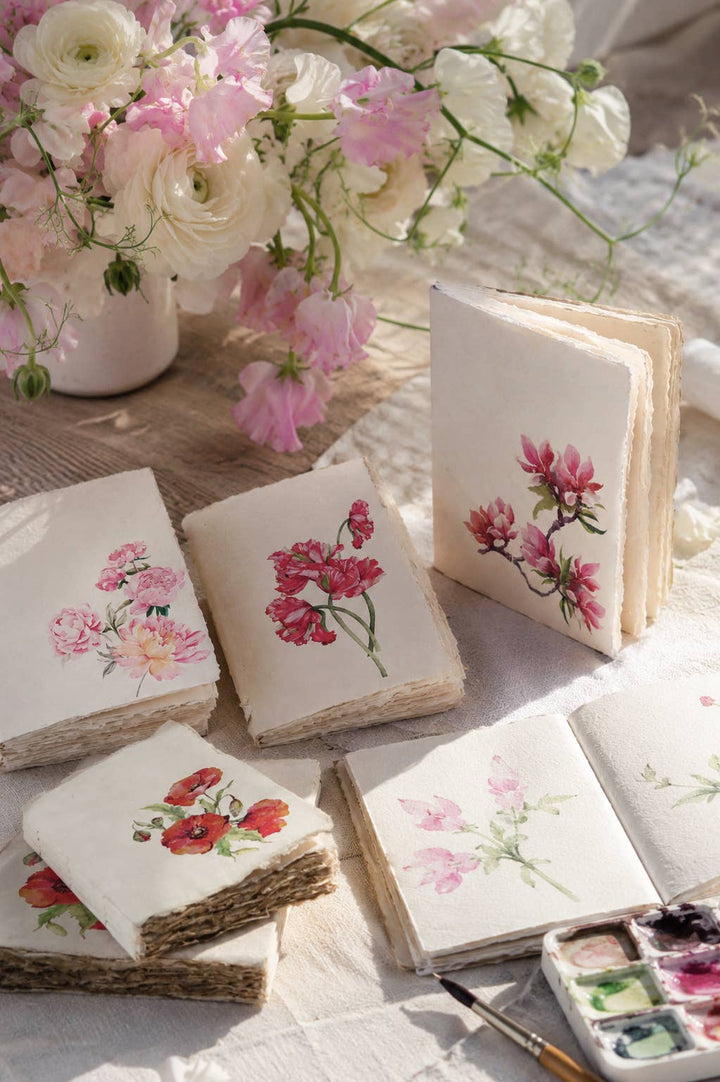Kukkakuvioisia muistikirjoja pöydällä kukkien ja vesiväripalettien vieressä; yhdessä kirjassa näkyvät avoimet paperisivut. / Floral notebooks on a table beside flowers and watercolour palettes, with one notebook open to show its paper pages.